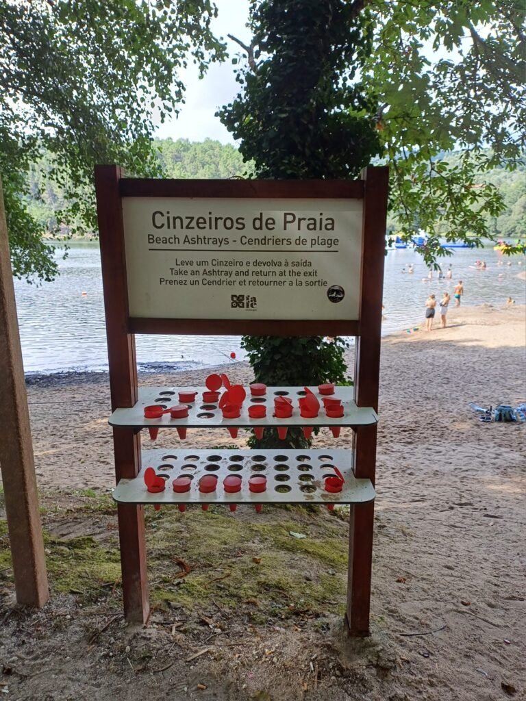 A stand with a sign for borrowing portable ashtrays at Barragem da Queimadela, encouraging visitors to keep the area clean by returning them after use.